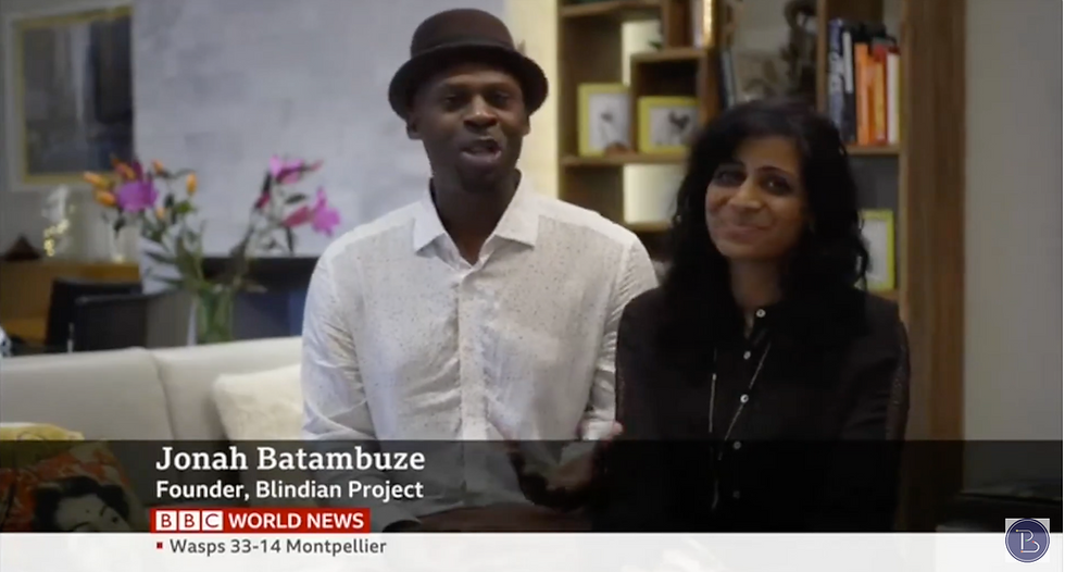 Jonah Batambuze, founder of the Blindian Project, and his partner speaking during a BBC World News interview about Black and South Asian relationships, seated together on a sofa with a warm, lived-in background.