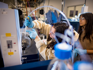A woman in goggles and gloves uses a pipette in a lab, with a colleague observing. Equipment and tubes fill the background.