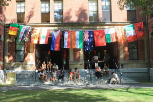 Students sit on steps of a building adorned with international flags. Sunny day, brick backdrop, relaxed mood.