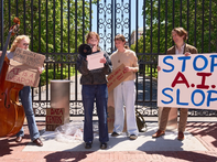 Protesters with signs against AI stand by a wrought-iron gate, one speaks into a megaphone.