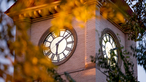 Brick clock tower with twin Roman numeral clocks seen through blurred yellow leaves in warm evening light on Whitman's college campus.