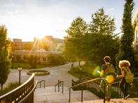 Two students with backpacks walk down campus steps at sunset, with sun flare, trees, buildings, and a courtyard fountain.