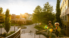 Two students with backpacks walk down campus steps at sunset, with sun flare, trees, buildings, and a courtyard fountain.