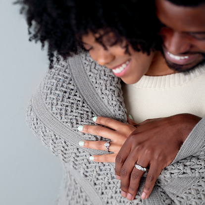 A man hugging a woman from behing, both are smiling ear to ear wearing white gold and diamond rings.
