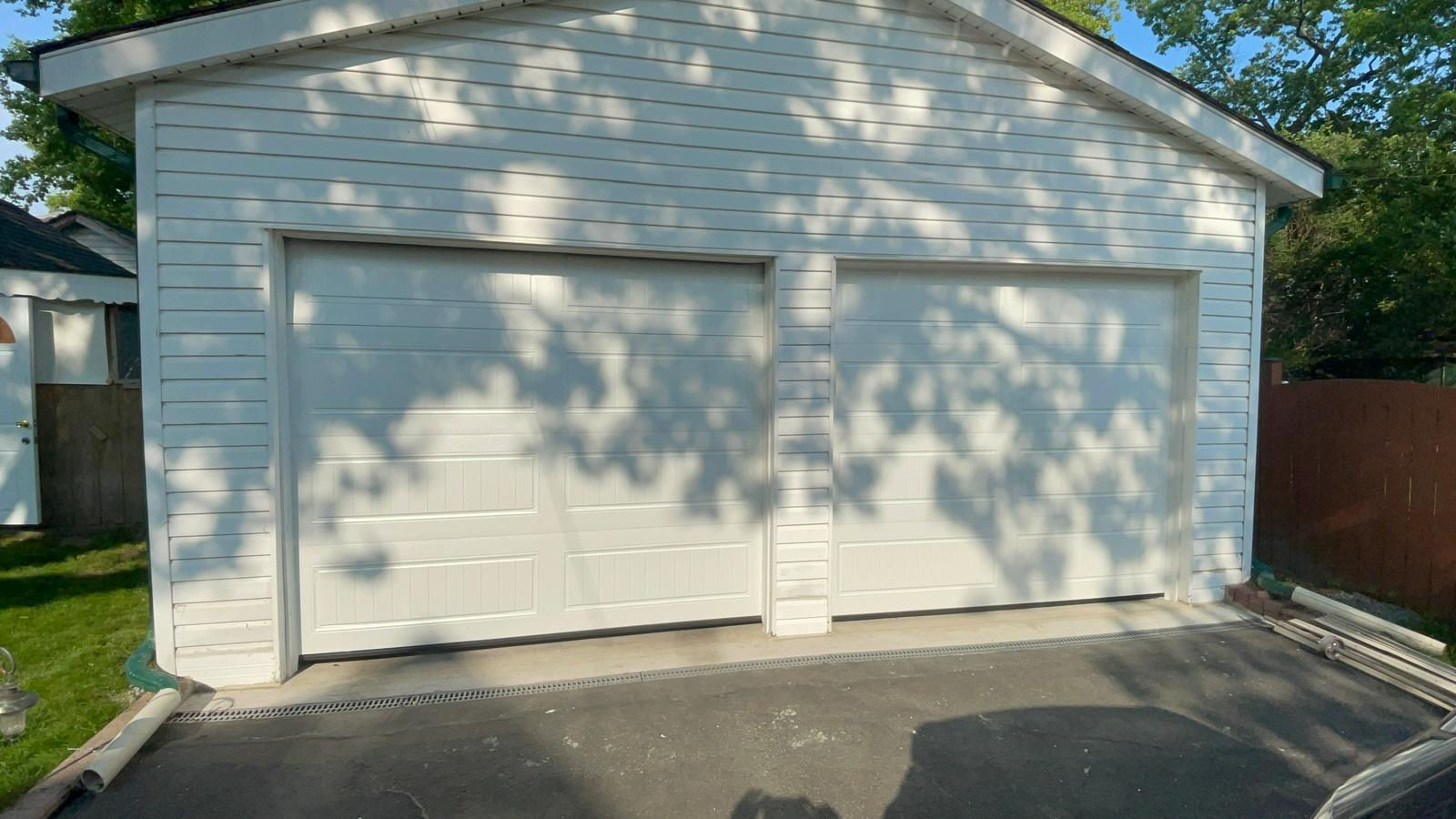 White garage doors, asphalt driveway, shadows, trees, and bright daytime shot. Rh Garage Door Repair