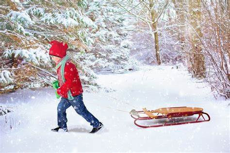 Child in red coat and hat pulls wooden sled through snowy forest. Snow-covered trees in background create a cheerful winter scene.