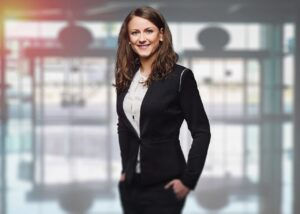 Smiling woman in a black suit stands confidently in a bright, blurred office setting. Light filters through large windows, creating a warm ambiance.