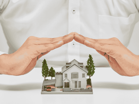 Duas mãos formando cobertura sobre uma casa em miniatura com árvores, conceito de proteção do patrimônio.