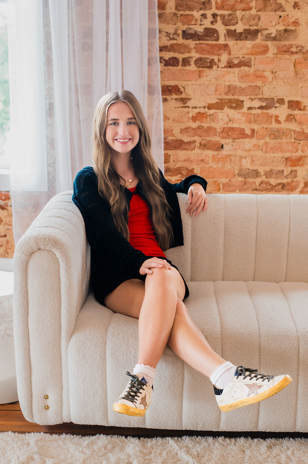The image shows a young woman with long, light brown hair seated comfortably on a textured, off-white couch. She is smiling at the camera and wearing a short, red garment, possibly a dress or romper, under a dark, possibly navy or black, open-knit cardigan. Her legs are crossed, and she is wearing white socks and light-colored sneakers with a star on the side. Behind her, a large window is visible on the left with sheer curtains, and a textured, exposed red brick wall is on the right.