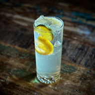Highball glass sitting on distressed wood table with clear cocktail and floating muddled lemon and lime. Menu out of focus to the left