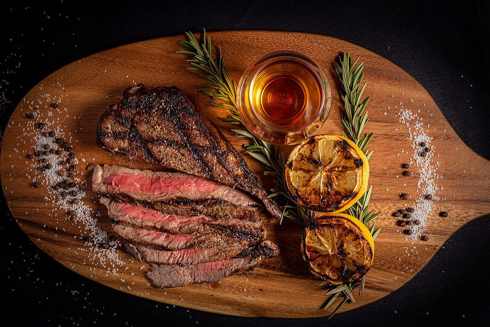 Top view looking down onto cutting board with steak cooked medium and sliced. Grilled lemons and rosemary on the side with salt and peppercorns sprinkled about. Glass of whiskey served neat.