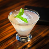 Stemless martini glass on copper bar and menu sitting in background. Slightly off-yellow colored cocktail with one large ice cube and two large leaves of fresh mint floating on top.