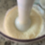 Blender immersed in a clear narrow container filled with oil and eggyolk getting blended. With lemon slices on a kitchen countertop. Nearby are a bowl with blue patterns and a plate with boiled eggs.