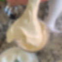 A spoon scoops homemade mayonnaise from a transparent container on a speckled countertop. Boiled egg halves and a white blender are nearby.