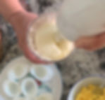 Hands blending creamy mixture in a bowl on a marble countertop. Nearby are halved boiled eggs on a plate and mashed yolks in a bowl.