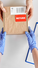 Gloved hands pass a cardboard box labeled RETURN with barcode over a clipboard and pen on a white worktable.