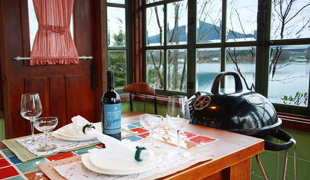 Dining table with barbecue grill next to it and window overlooking water and mountain