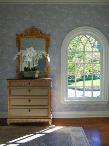 Classic bedroom vignette with antique dresser, arched window, and floral wallpaper by Design by Shelley Mcilroy Design.