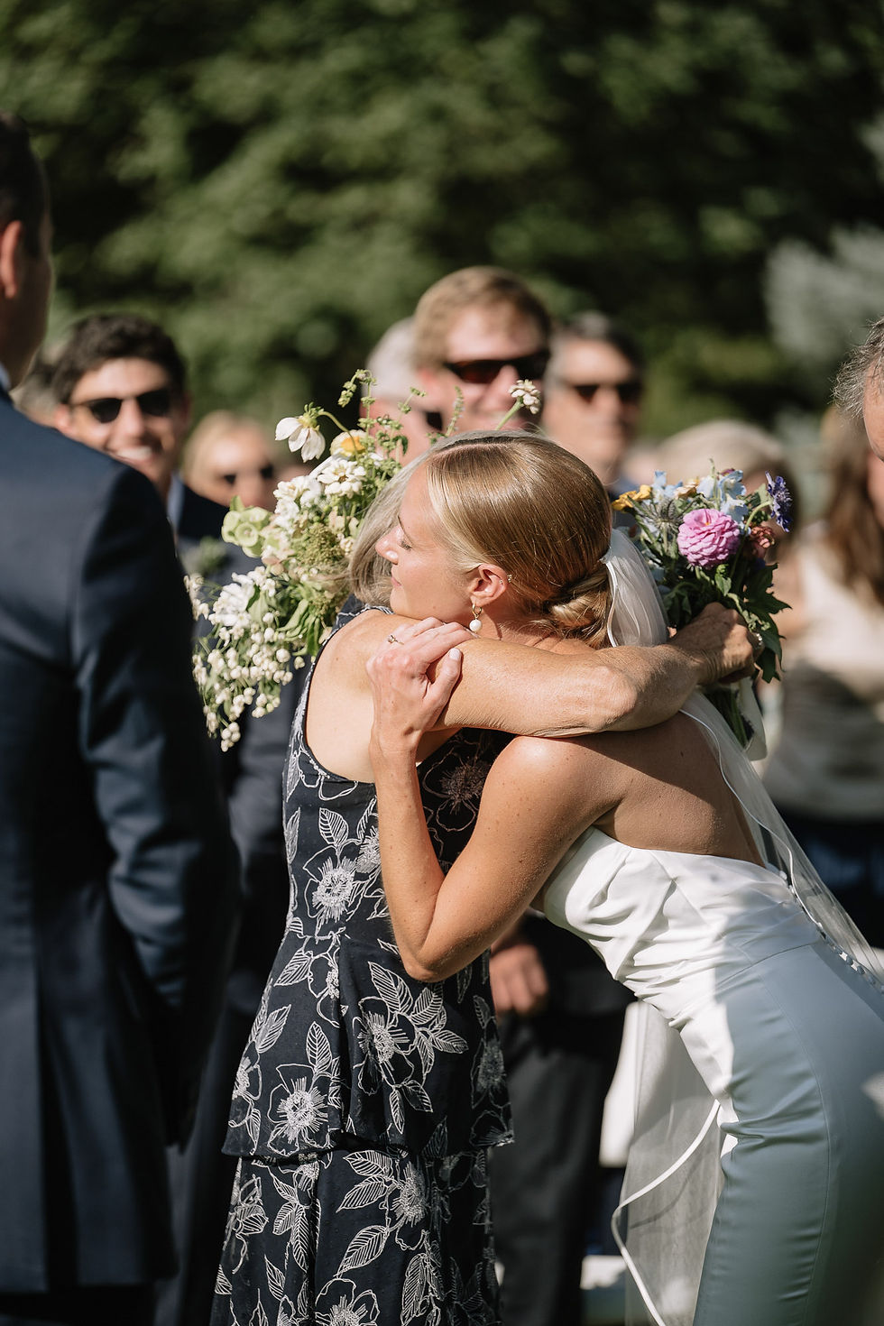 Bride in white dress hugs a guest in a black floral dress at an outdoor wedding, with guests and bouquets in sunlight.