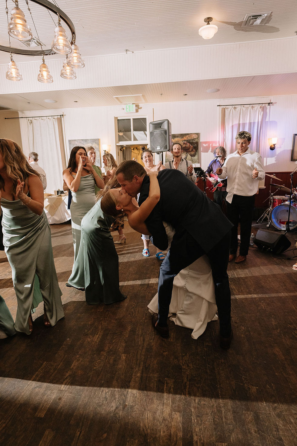 Wedding reception with a couple kissing on the dance floor while guests clap; band plays in a bright hall.