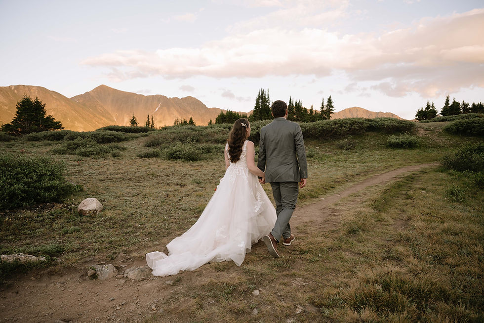 Bride in flowing white gown and groom in gray suit walk hand in hand on a mountain trail at sunset, with pine trees and peaks behind near Keystone, CO.
