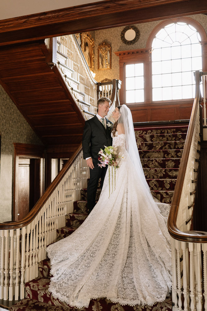 Bride and groom in wedding attire on a grand staircase in the Stanley Hotel, smiling at each other; lace gown and bouquet.