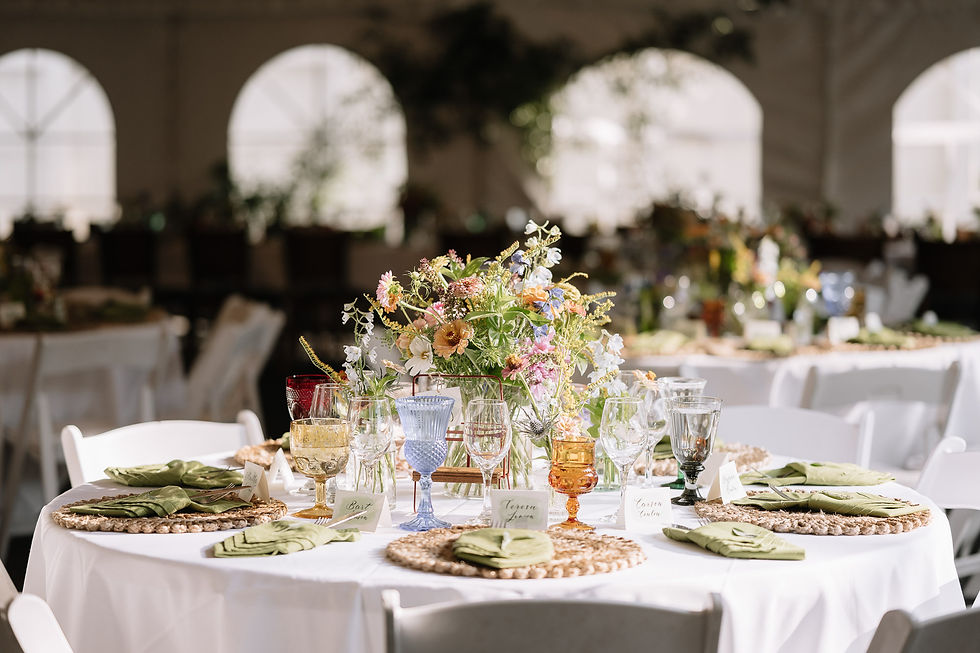 Elegant banquet table with pastel floral centerpiece, colorful glassware, green napkins, and place cards in a sunlit hall with arched windows