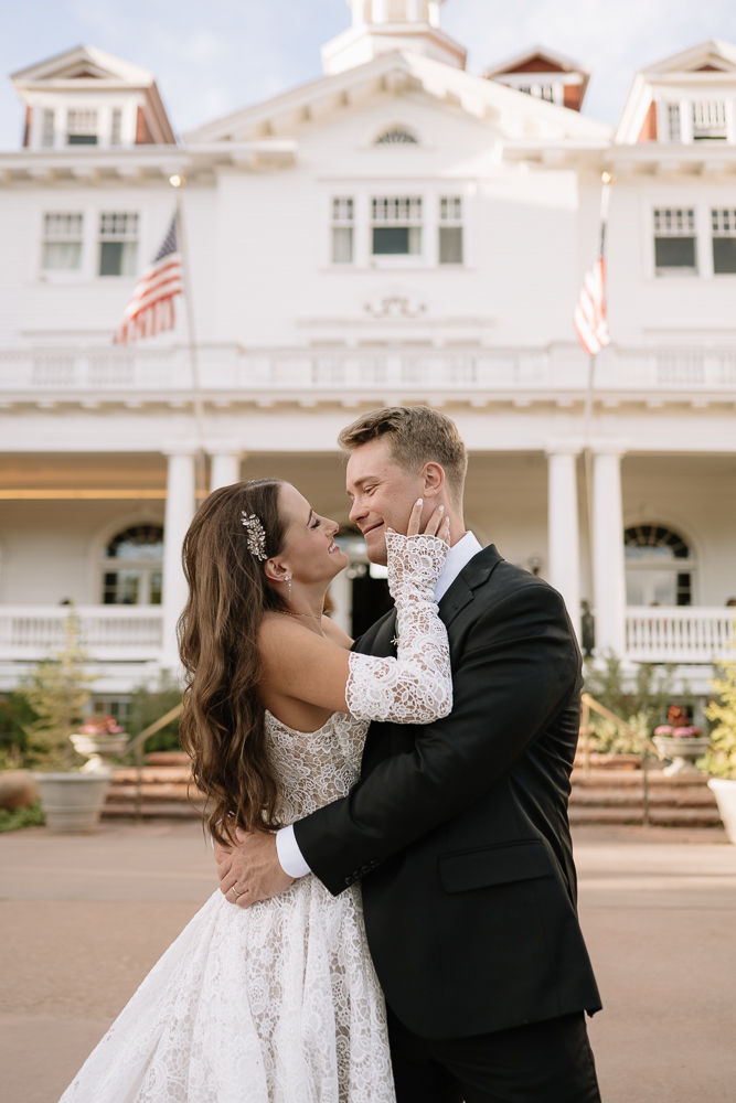 Bride in lace gown and groom in black suit embrace and smile outside the Stanley Hotel with American flags.