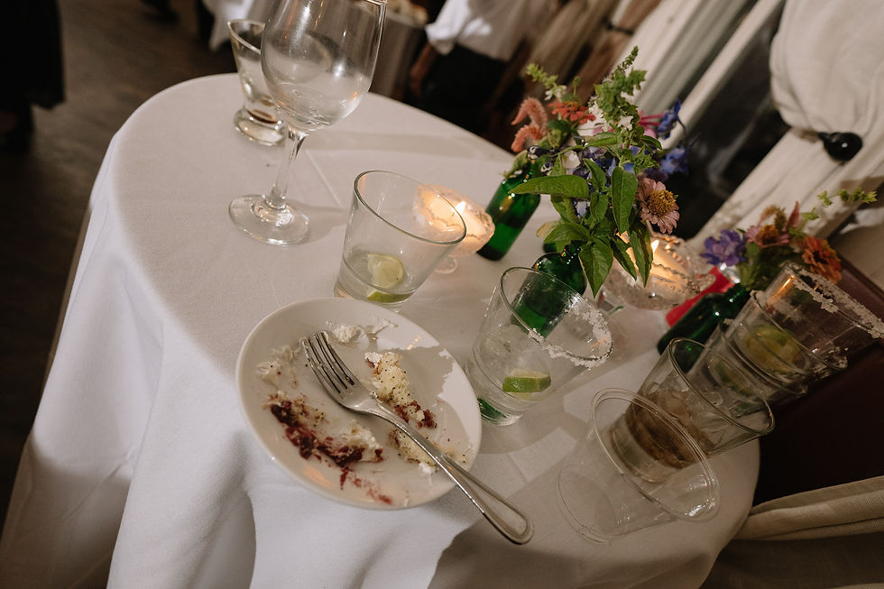 Partly eaten dessert on a white-clothed banquet table with empty glasses, candles, and colorful flowers at a softly lit reception