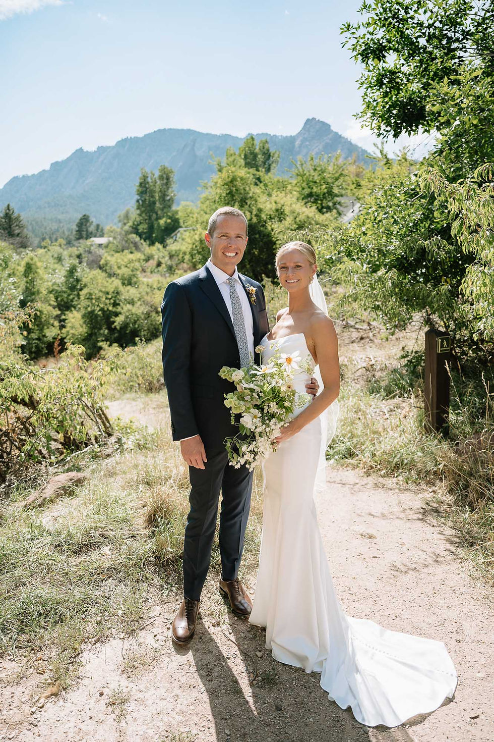Smiling bride and groom in wedding attire pose on a sunlit mountain trail, holding a bouquet with green trees behind them.