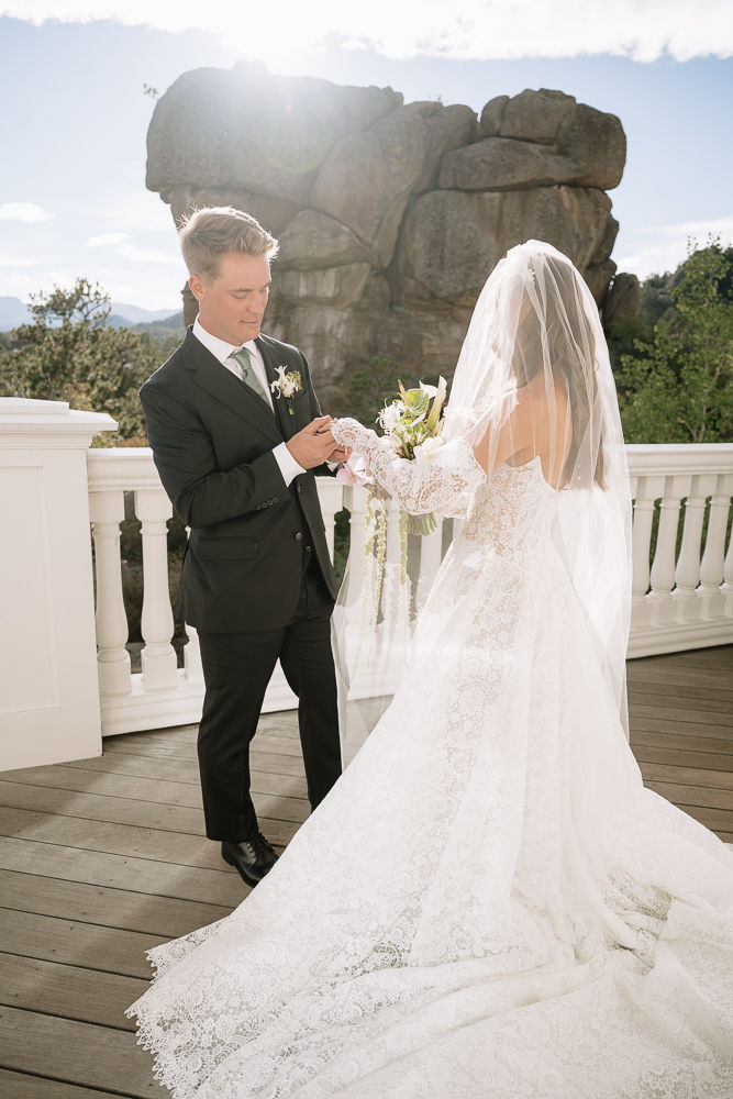 Groom in black suit places a ring on bride in lace veil and gown on a sunlit terrace, with a rock formation behind.