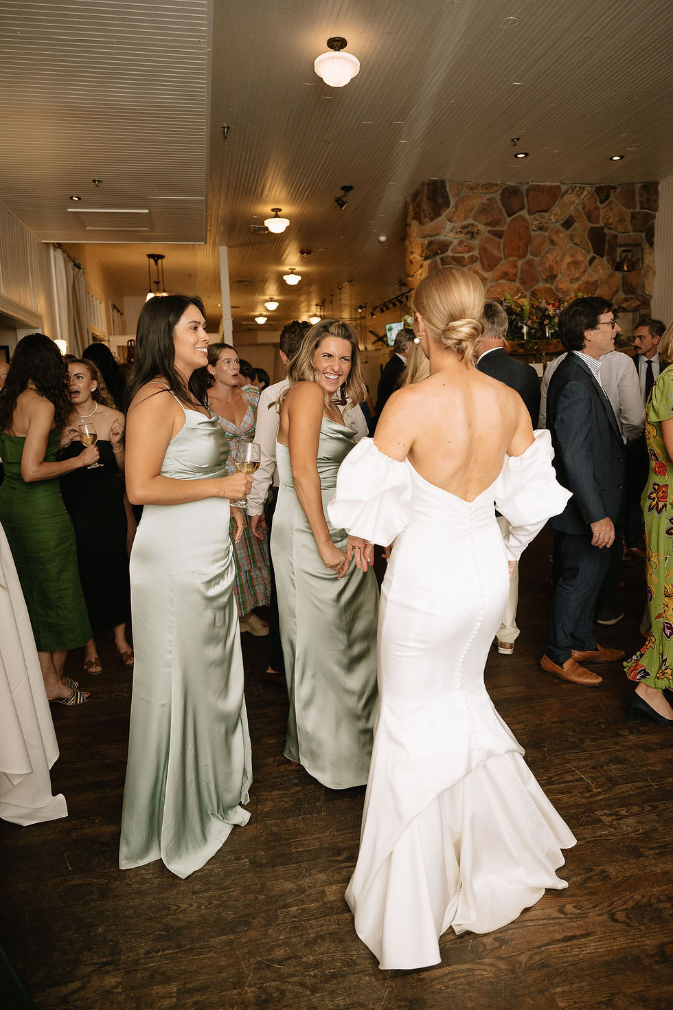 Guests in satin green and white gowns chat at an indoor wedding reception, smiling under warm lights on a wood floor.