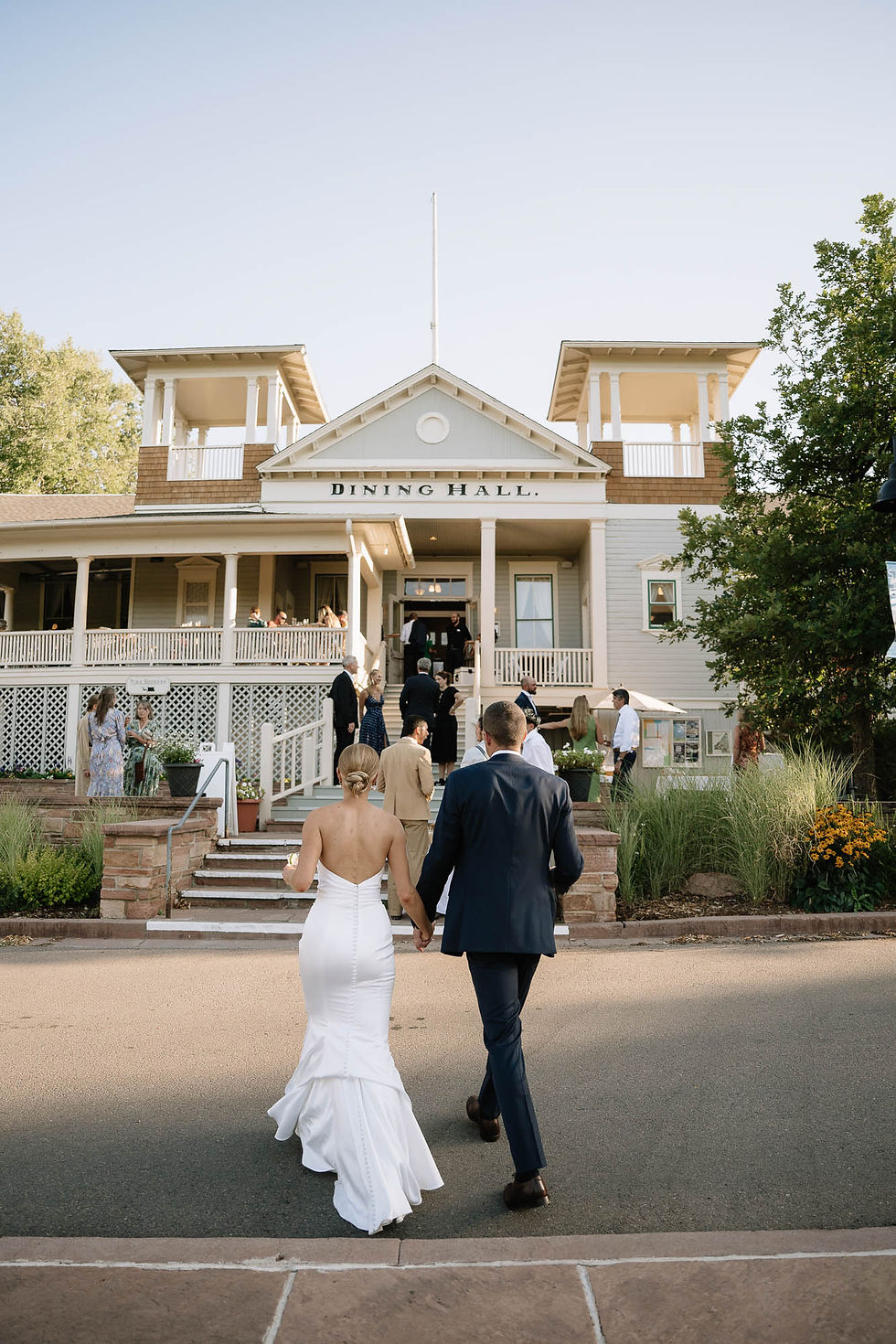 Bride in white gown and groom in navy suit walk toward a Dining Hall at an outdoor wedding, with guests gathered nearby.