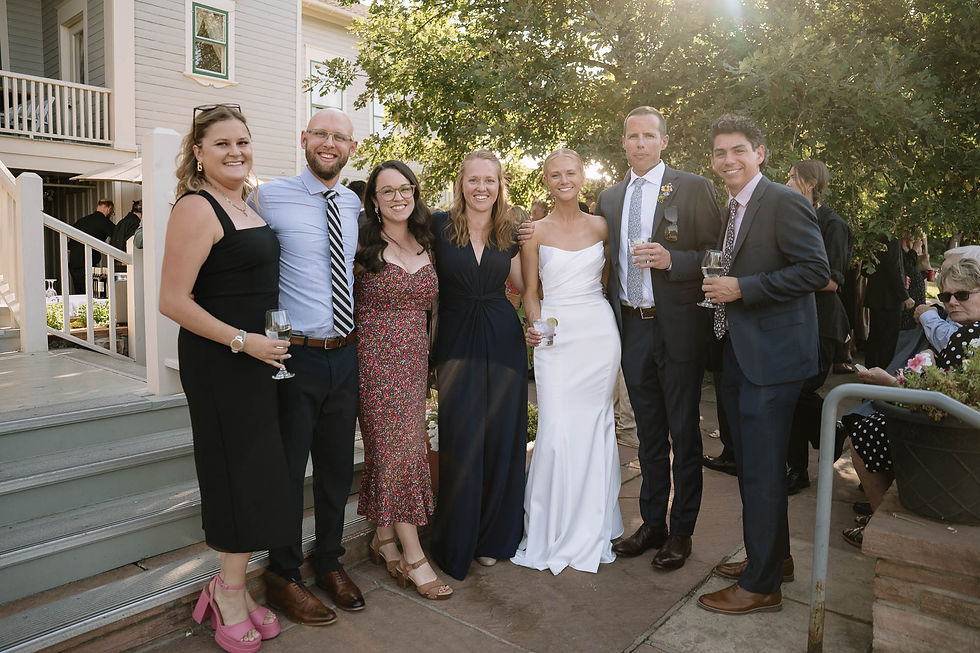 Smiling wedding guests pose outdoors by a house, holding wine glasses in formal attire under trees in warm sunlight.