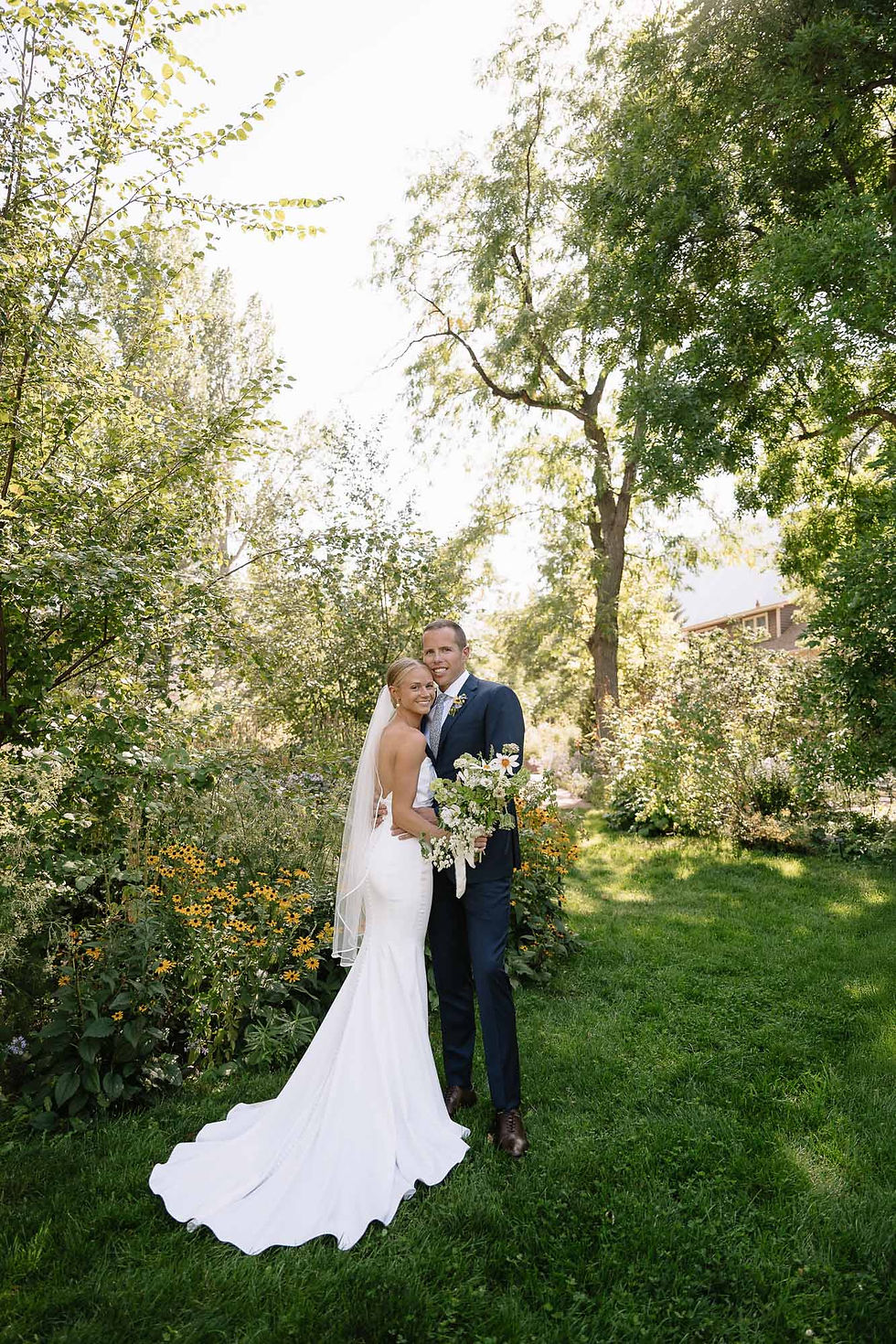 Bride and groom pose smiling in a sunny garden, bride in white gown and veil, groom in navy suit holding bouquet at Chautauqua.