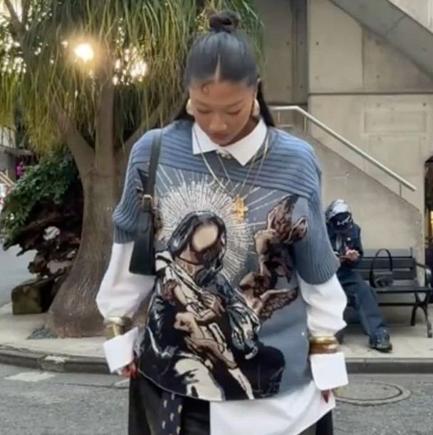 Urban streetwear outfit with oversized graphic sweater layered over white shirt and wide-leg black pants, mixing casual tailoring, contemporary fashion trends, and everyday luxury styling in city setting.