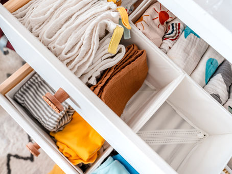 A white nursery drawer divided into compartments, holding folded baby blankets, muslin cloths, and colorful baby clothes. The drawer is neatly arranged with soft fabrics and vibrant colors.