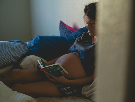 Pregnant woman reading a children’s book in bed, surrounded by pillows, showing a calm and cozy prenatal moment.