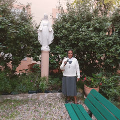 Soeur Georgette est debout à côté d'une statue de la vierge. Elle tient un micro dans les mains et regarde la caméra.