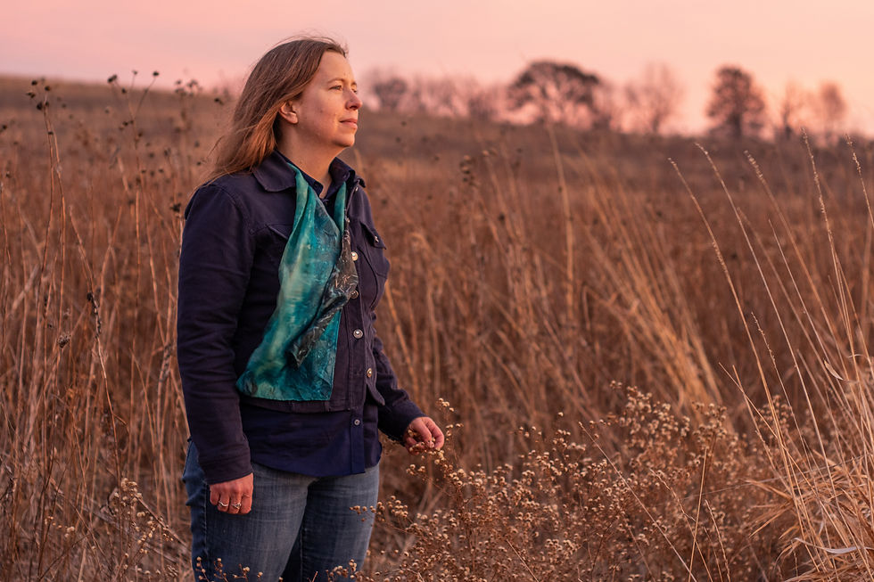 Kaia Fowler stands in a field of prairie grass in autumn looking off into the distance, touching one a dried flower in front of her.