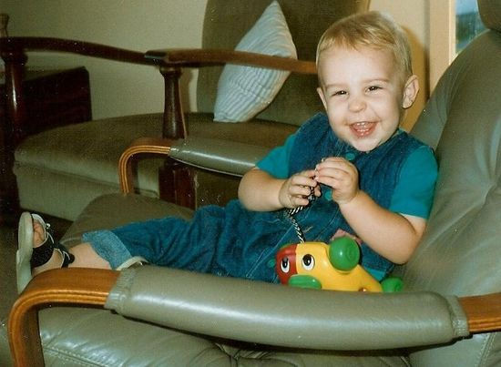 A cheerful toddler in a blue jumpsuit laughs while holding a toy caterpillar in a vintage brown armchair.