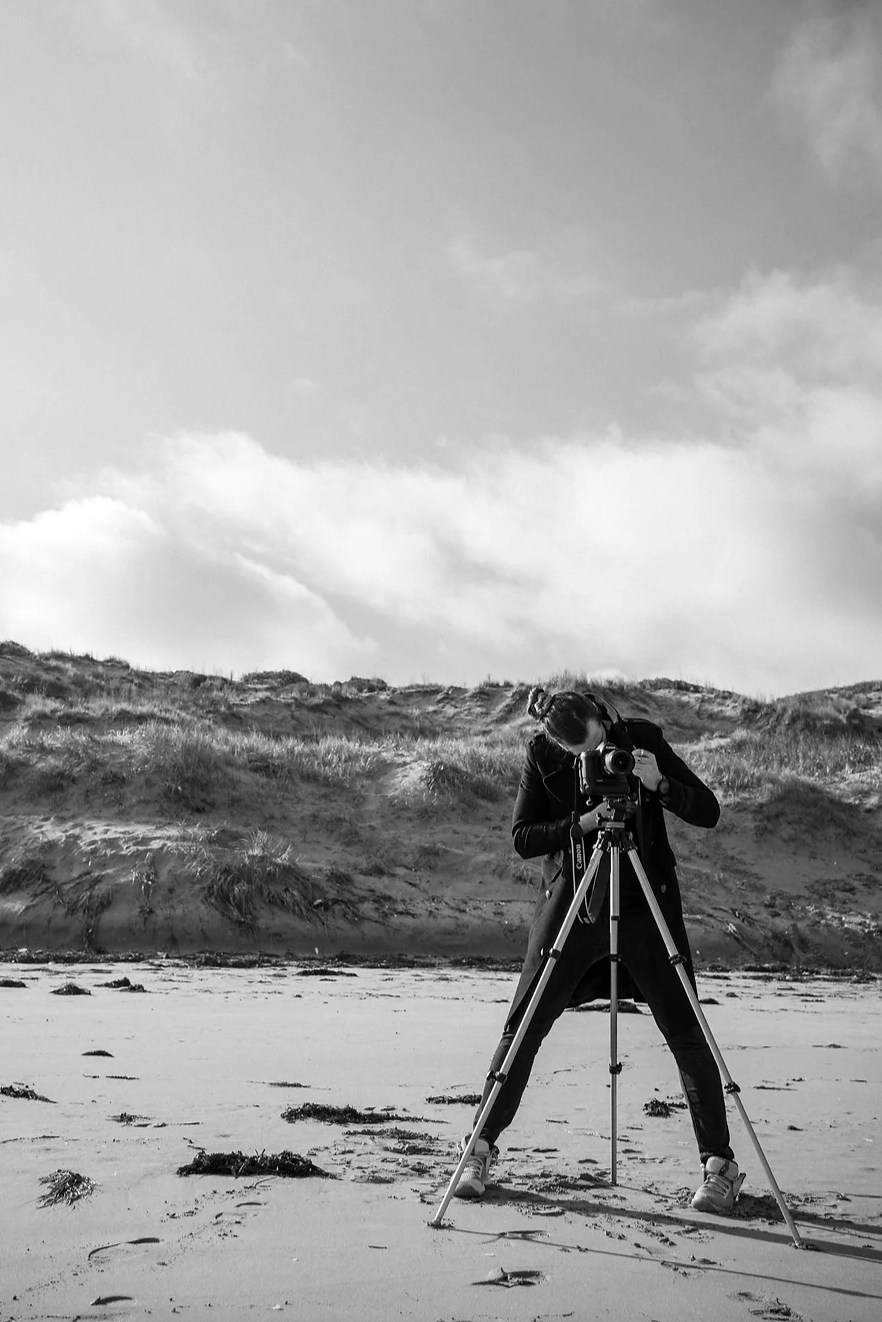 A photographer crouches behind a camera mounted on a tripod, aiming at a scenic beach with grassy dunes under a cloudy sky