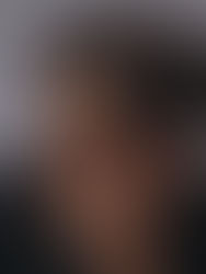 Curly-haired person with rings and tattoos holds fingers near eyes, gazing thoughtfully. Neutral background, moody lighting.