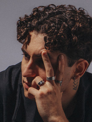 Curly-haired person with rings and tattoos holds fingers near eyes, gazing thoughtfully. Neutral background, moody lighting.