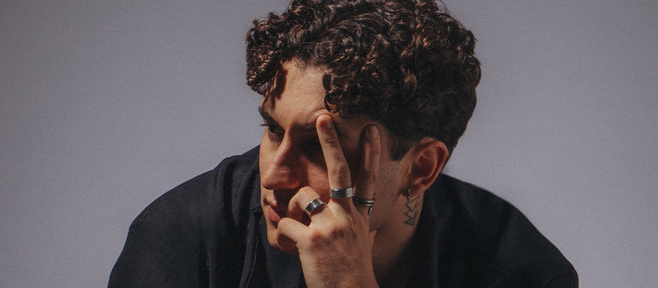 Curly-haired person with rings and tattoos holds fingers near eyes, gazing thoughtfully. Neutral background, moody lighting.