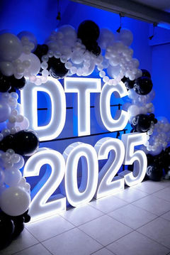 White glowing 'DTC 2025' sign with balloon arch against blue wall.