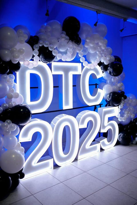 White glowing 'DTC 2025' sign with balloon arch against blue wall.