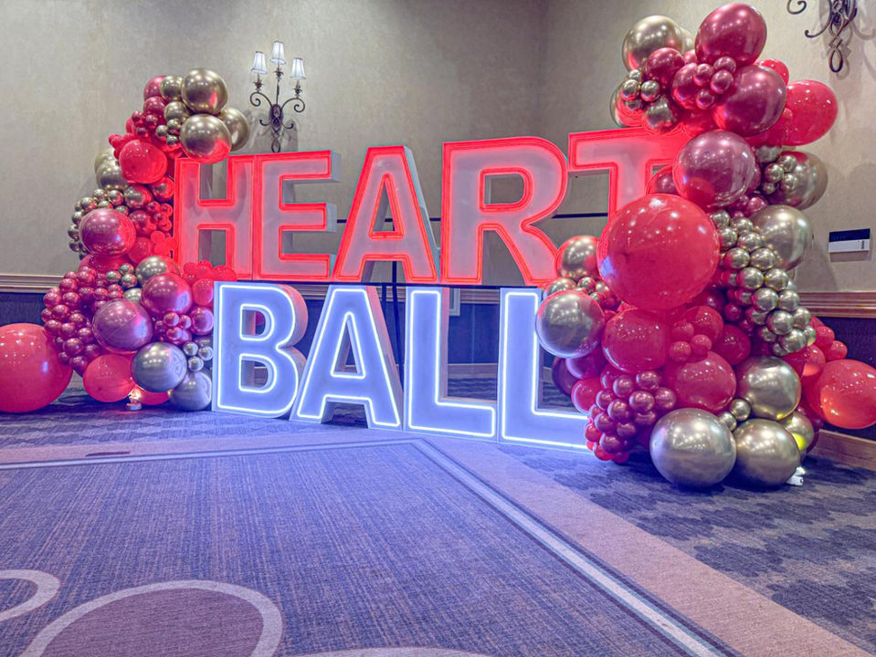Illuminated HEART BALL sign with glowing letters, surrounded by red and gold balloons.