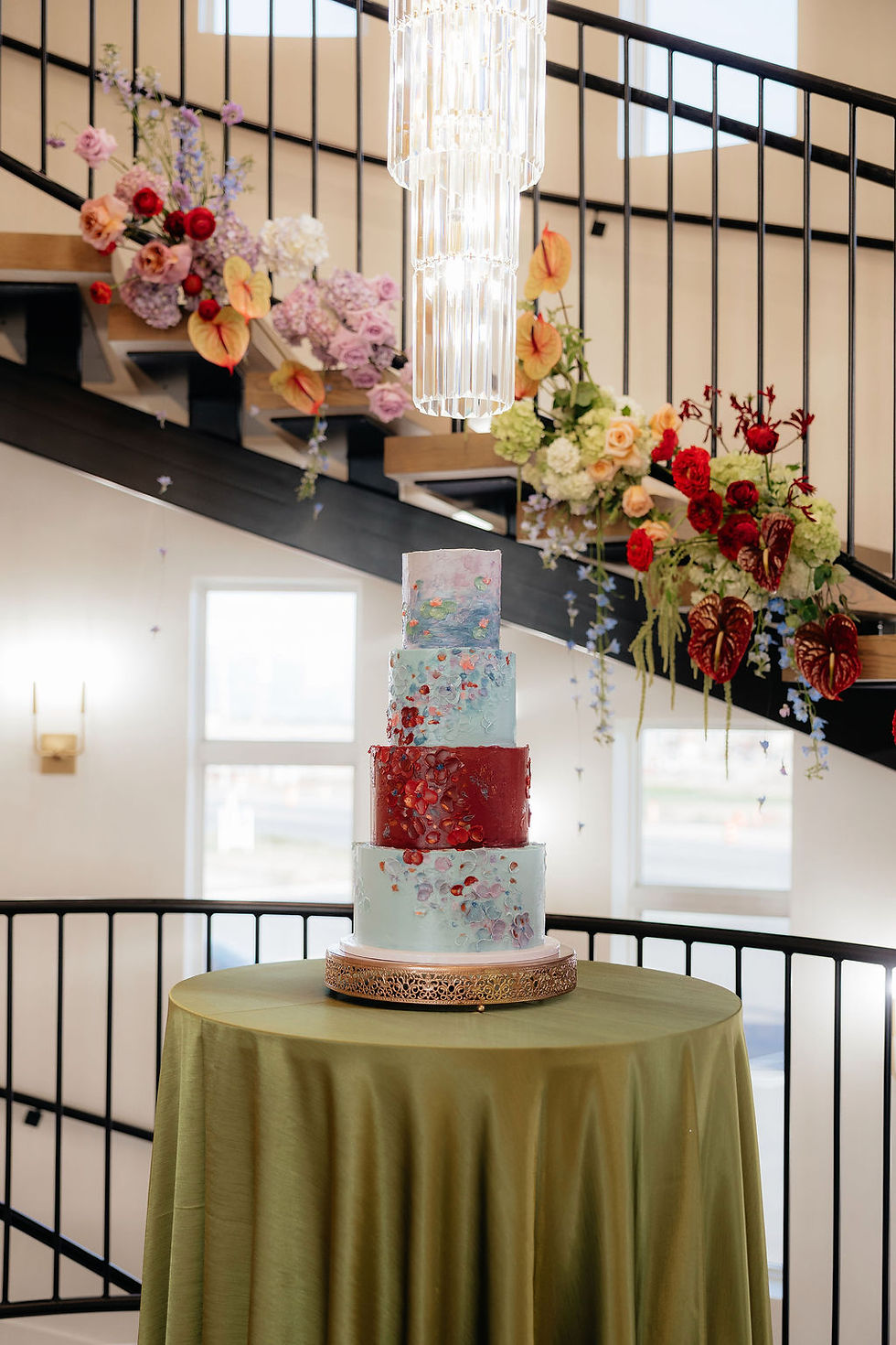 Tiered cake with colorful patterns on a green table, under a chandelier. Staircase with flowers in the background, creating an elegant setting.