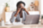 Fatigued female wearing glasses pushing glasses up holding eyes. Female is sitting at a desk in front of an open laptop. There is a coffee cup, plant, paper, and pens still on her desk. The background is blurry but there is a shelf with a plant, lamp, and decor.